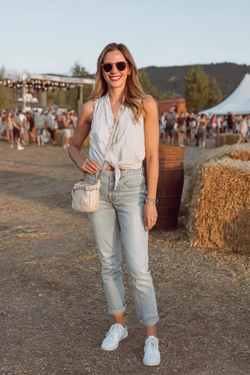 A woman in light-wash jeans with a sleeveless tie-front blouse and white sneakers, accessorized with sunglasses and a small crossbody bag