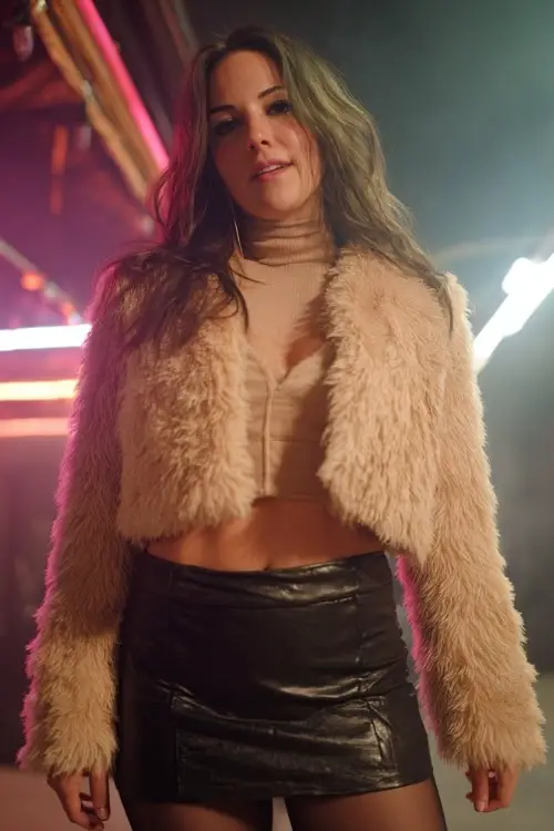 A curvy woman wears a faux fur cropped jacket over a turtleneck top and leather mini skirt, styled with tights and cowboy boots, standing in a night concert setting with neon lights 