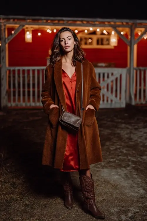A woman wears a floor-length wool coat layered over a satin slip dress, styled with tall cowboy boots and a leather crossbody bag 