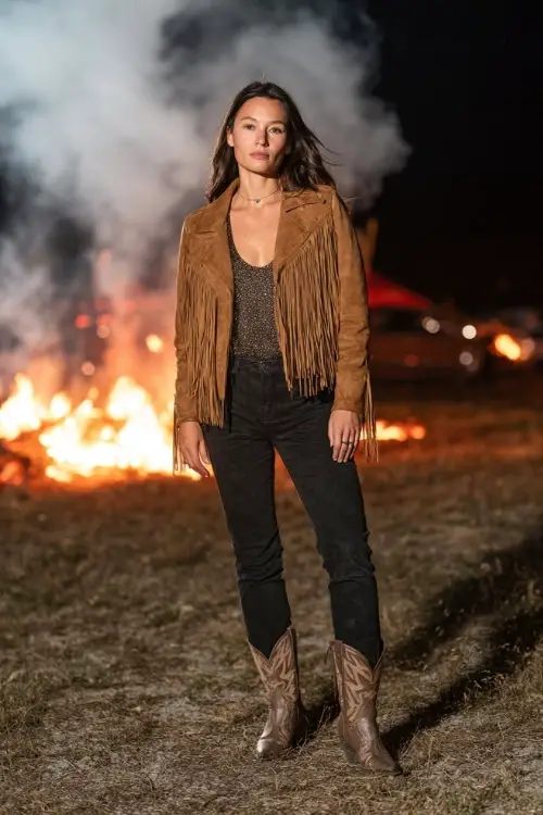A woman wears a fringe suede jacket over a sparkly tank top and black jeans, styled with pointed-toe cowboy boots, standing near a bonfire at a nighttime country festival 