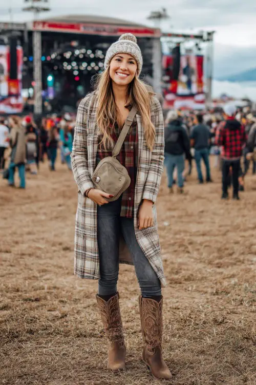 A woman wears a long plaid coat with skinny jeans, suede cowboy boots, and a crossbody bag, styled with a knit beanie at an outdoor music festival stage 