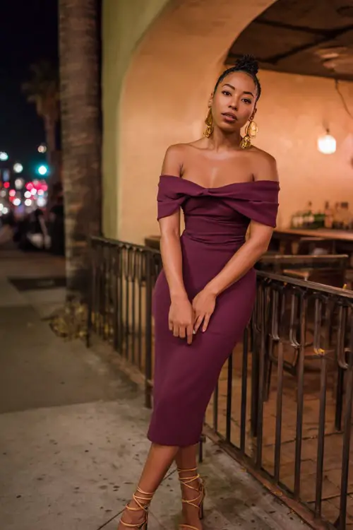 A Black woman in a chic off-the-shoulder plum midi dress with strappy heels and statement earrings, posing outside a lively fall night bar, full body outfit visible 