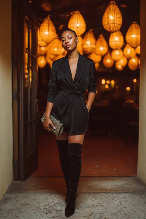 A Black woman in a sleek black blazer dress with thigh-high boots and a clutch bag, standing at the entrance of a chic fall cocktail lounge with glowing lanterns 