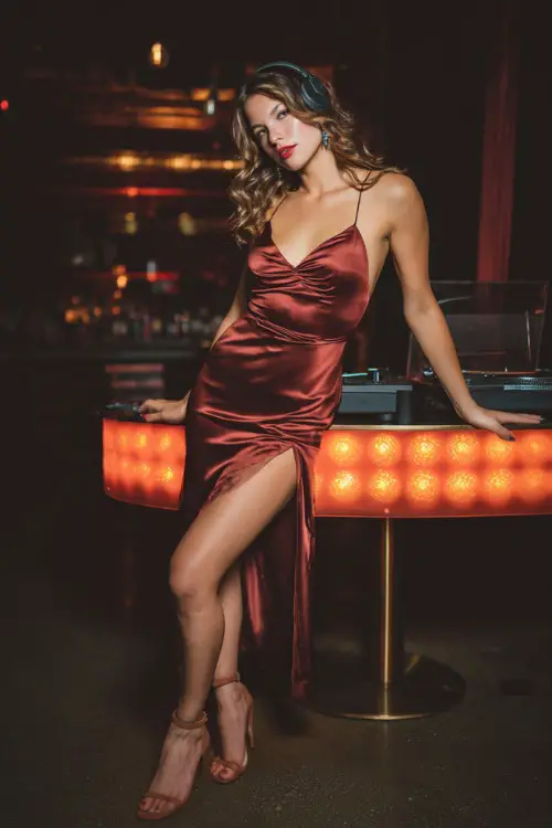 A woman in a fitted burgundy satin slip dress with a thigh slit, paired with ankle strap heels, posing near a glowing DJ booth in a fall nightclub 