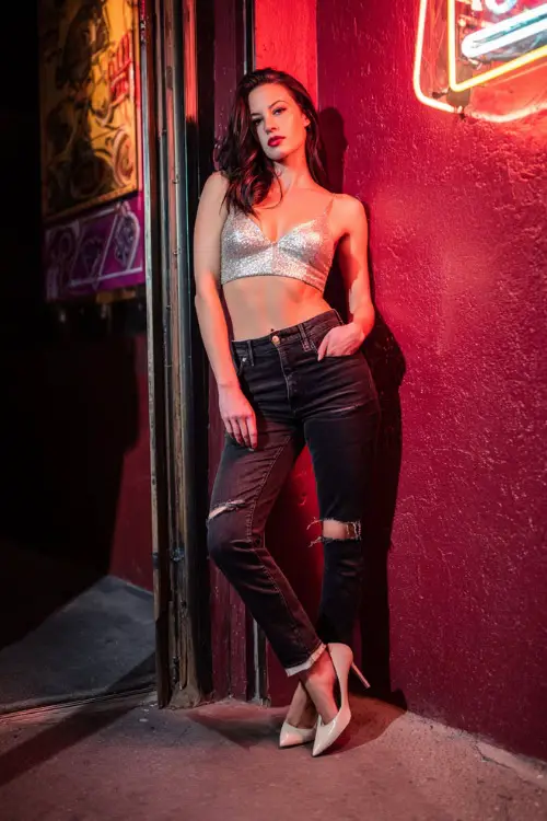 A woman in ripped black skinny jeans, a sequined silver top, and stiletto heels, posing near a neon-lit nightclub entrance, full body outfit visible