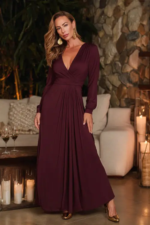 A woman over 40 in a deep wine-colored wrap maxi dress with long sleeves, paired with gold heels and statement earrings, posing elegantly inside a candlelit autumn lounge
