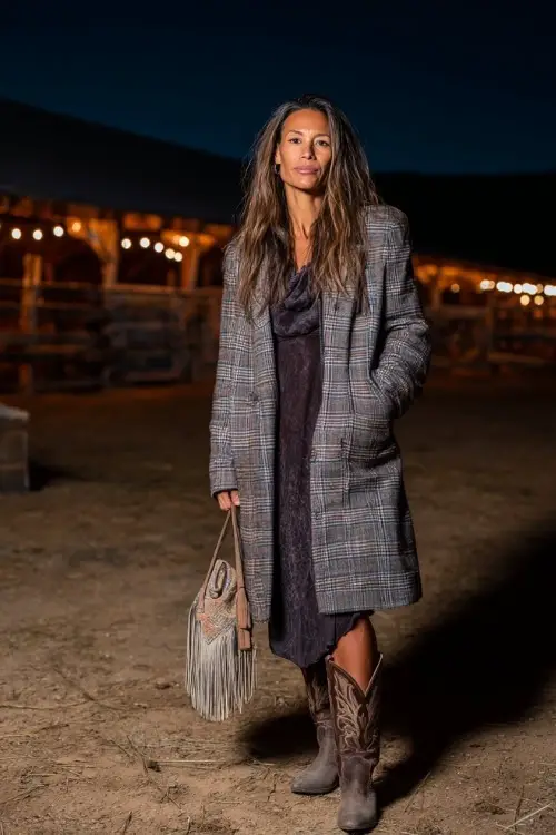 A woman over 40 wears a long plaid coat over a velvet dress with cowboy boots, holding a fringe bag at a nighttime barn concert 