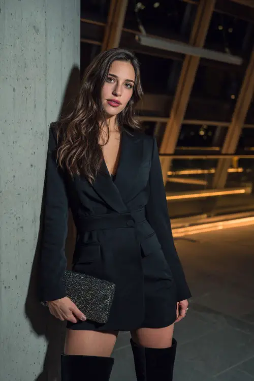 A woman wearing a tailored black blazer mini dress with long sleeves, paired with over-the-knee boots and a glittery clutch 