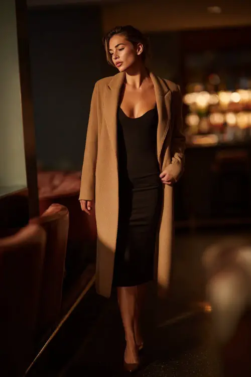 A woman wearing a tailored camel coat draped over a fitted midi dress with heels, walking into a candlelit fall lounge 