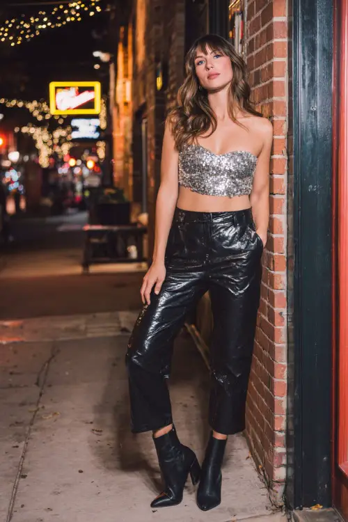 A woman wearing black faux leather pants, a sequined silver crop top, and heeled ankle boots, posing at the entrance of a busy casual bar on a crisp fall night