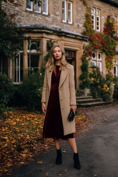 A woman wears a burgundy velvet midi dress with long sleeves, layered under a tailored camel wool coat, styled with black heeled boots and a clutch