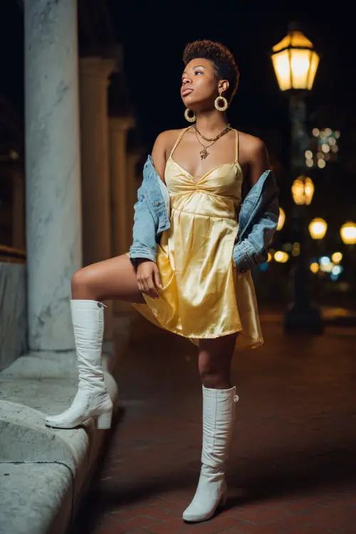 A woman wears a gold satin dress paired with a denim jacket, hoop earrings, and tall white boots under the glow of concert lanterns