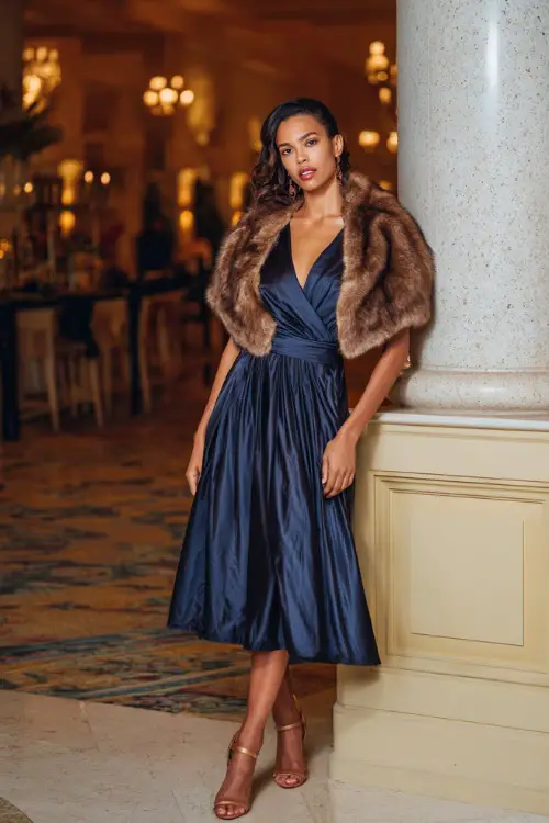 A woman wears a navy blue satin wrap dress with a pleated skirt, paired with a faux fur stole and ankle strap heels, posing at an elegant indoor ballroom wedding with warm golden lighting 