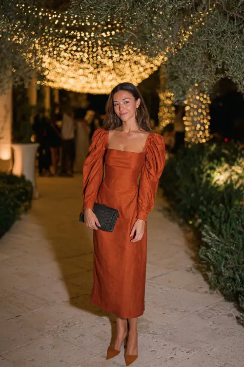 A woman wears a rust-orange midi dress with a square neckline and puff sleeves, styled with block heels and a clutch 