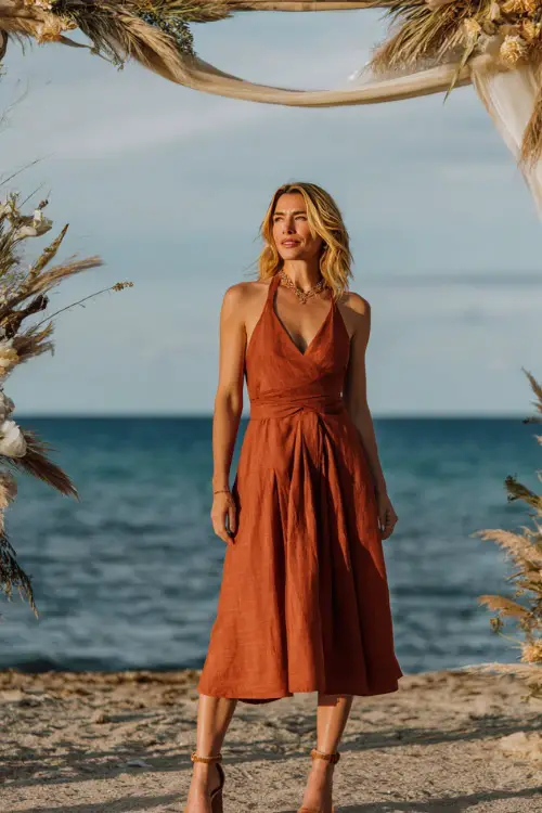 A woman wears a rust-orange sleeveless midi dress with a pleated skirt, paired with block heel sandals and minimal jewelry, standing under a boho wedding arch with the ocean horizon behind her
