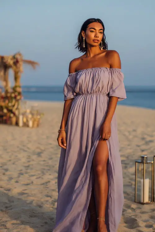 A woman wears a soft lavender off-the-shoulder maxi dress, styled with pearl sandals and drop earrings, posing barefoot on the sand at a romantic beach wedding with lanterns around 
