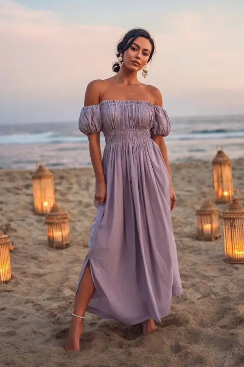 A woman wears a soft lavender off-the-shoulder maxi dress, styled with pearl sandals and drop earrings, posing barefoot on the sand at a romantic beach wedding with lanterns around
