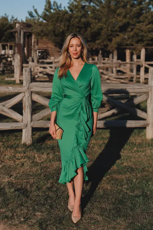 A woman wears a bright pine green midi wrap dress with a ruffled hem, styled with beige pumps and a small clutch 