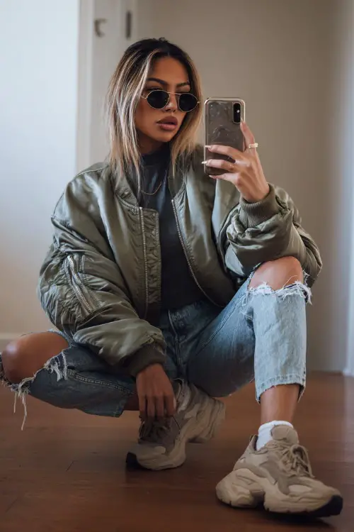 Baddie-inspired outfit with an oversized bomber jacket, ripped jeans, fitted turtleneck, and chunky sneakers, woman taking mirror selfie indoors with minimal background
