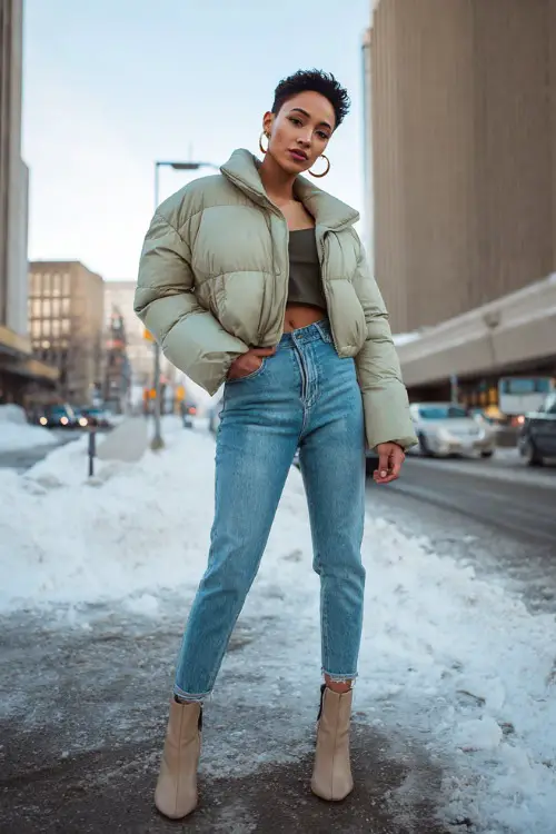 Confident woman in a cropped puffer jacket, high-waisted skinny jeans, heeled ankle boots, and hoop earrings, posing on a snowy city street with attitude 