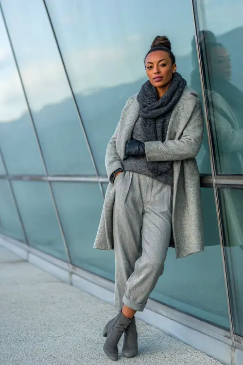 Sophisticated Black woman in a monochrome outfit with a gray peacoat, wool pants, gloves, and heeled boots, standing beside a glass building on a cold morning
