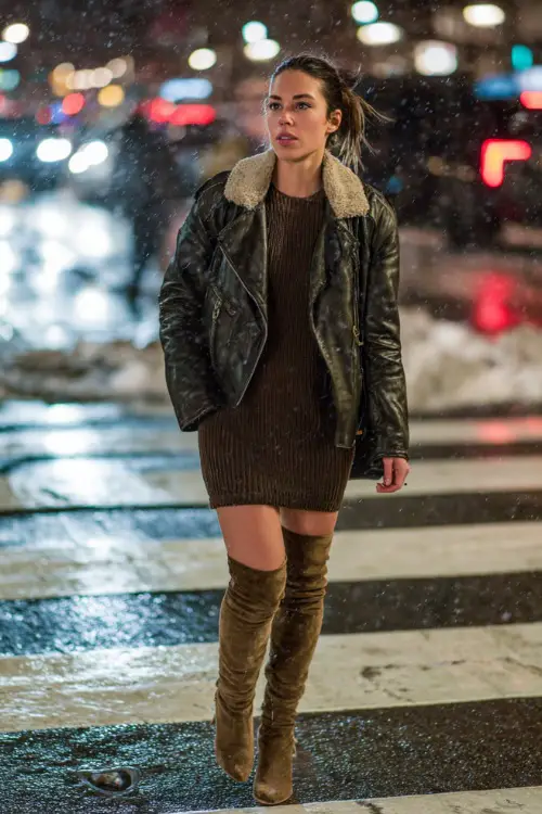 Trendy woman in a faux leather jacket, fitted knit dress, and thigh-high suede boots, walking through a snowy city crosswalk at night with lights reflecting off wet streets