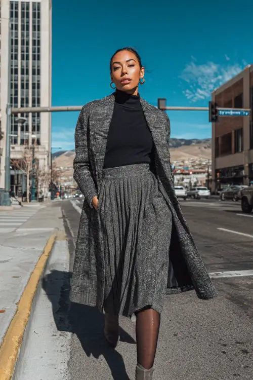 Woman in a tailored plaid coat, black turtleneck, pleated midi skirt, tights, and ankle boots, walking confidently across a city street on her way to work 