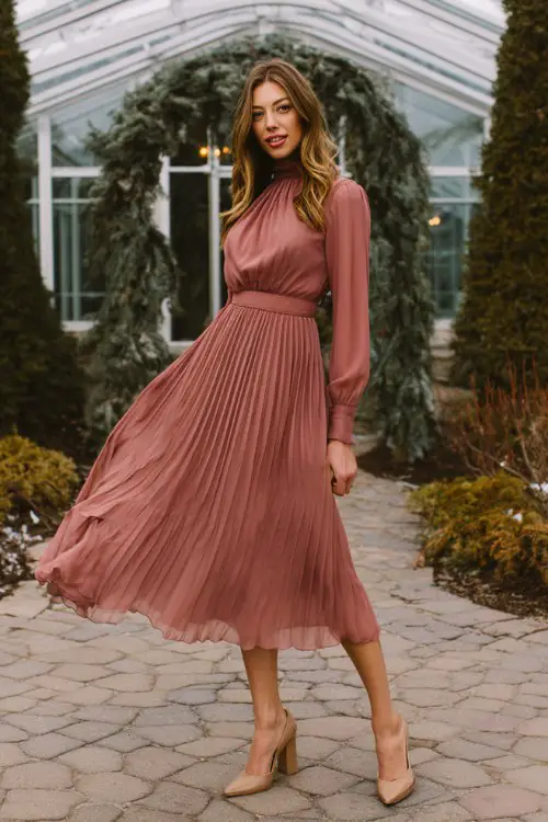 A woman wears a dusty rose long-sleeve tea-length dress with pleated chiffon and a high neckline, styled with block heels