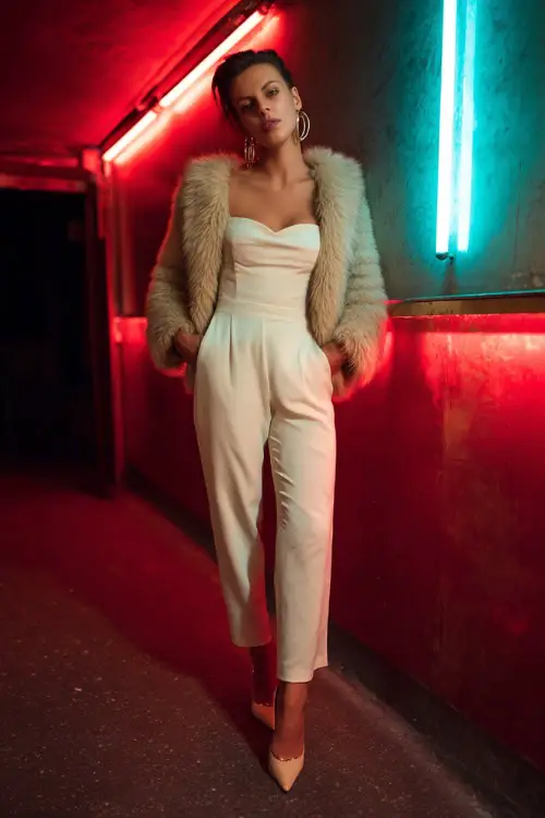 A bold woman wearing a sleek jumpsuit layered with a cropped faux fur coat, paired with pointed heels and hoop earrings, standing under neon club lights on a winter night