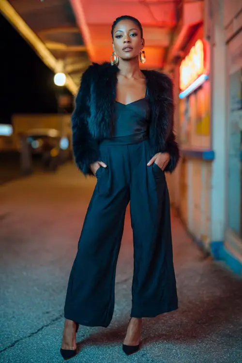 A bold woman wearing a sleek jumpsuit layered with a cropped faux fur coat, paired with pointed heels and hoop earrings