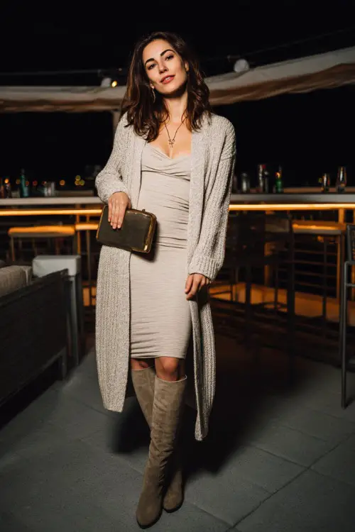 A chic woman wearing a long cardigan layered over a body-skimming midi dress, paired with tall boots and a statement clutch
