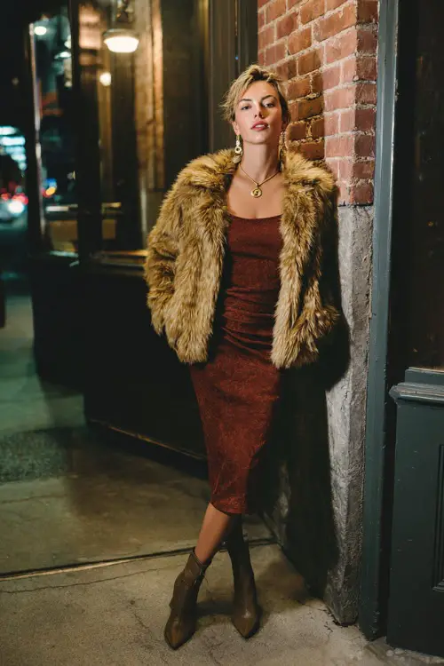 A chic woman wearing a short faux fur coat layered over a body-skimming midi dress, paired with ankle boots and bold earrings