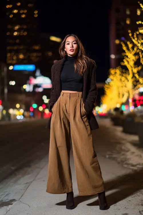 A confident woman wearing a fitted turtleneck tucked into high-waisted wide-leg trousers, layered with a cropped statement coat and platform heels 