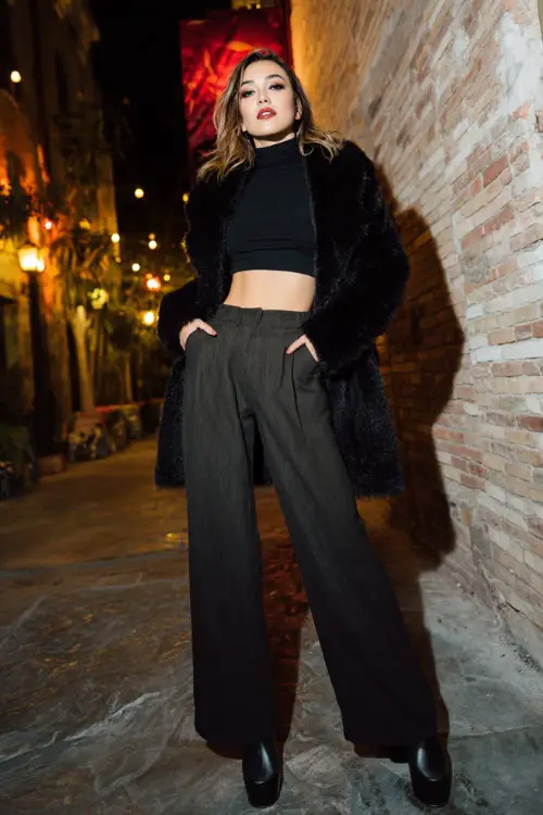 A confident woman wearing a fitted turtleneck tucked into high-waisted wide-leg trousers, layered with a cropped statement coat and platform heels 