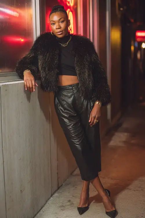 A fashionable Black woman in a cropped faux fur jacket worn over a sleek turtleneck top, paired with high-waisted leather pants and pointed-toe heels 