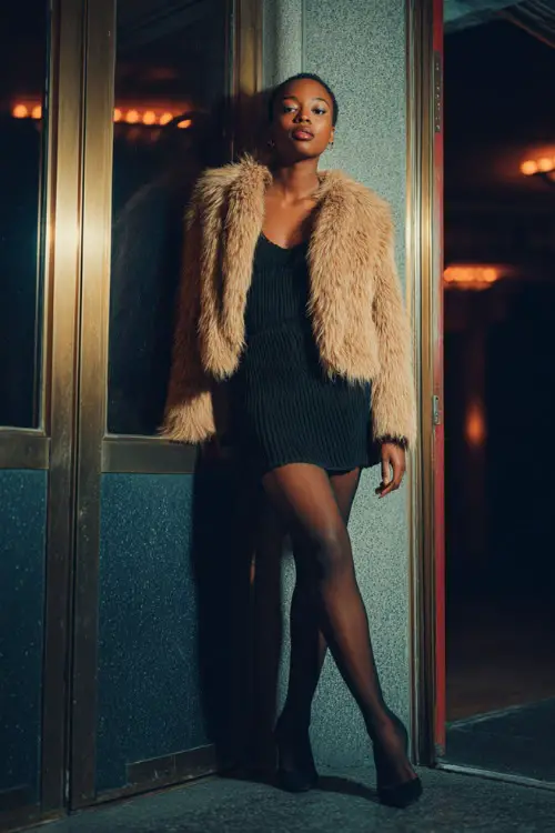 A stylish Black woman wearing a body-skimming knit mini dress layered with a short faux fur coat, styled with tights and pointed heels 