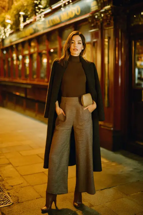 A stylish woman wearing a fitted long-sleeve top with wide-leg trousers, layered with a tailored coat and finished with heeled boots, accessorized with a clutch 