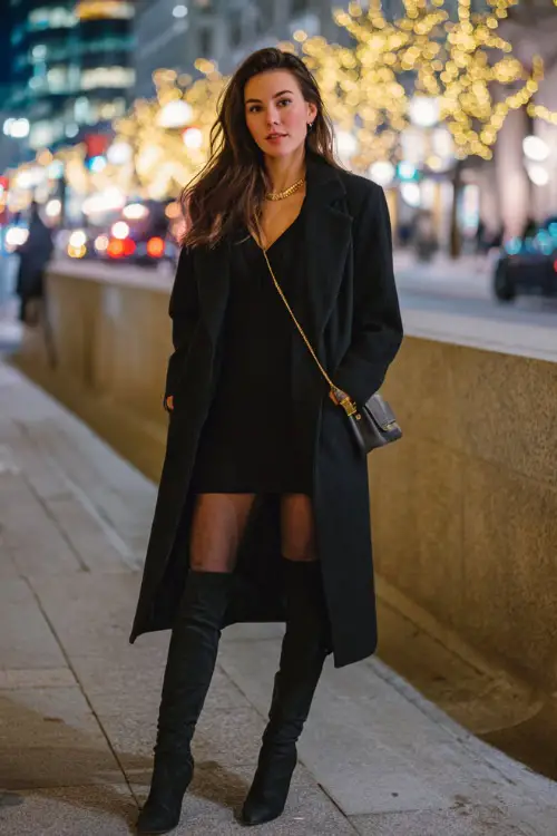 A stylish woman wearing a long black wool coat layered over a fitted knit dress, sheer tights, and knee-high leather boots