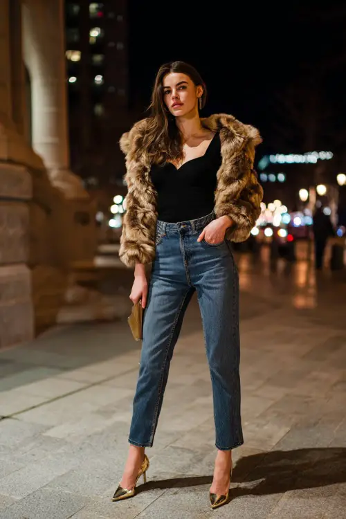 A stylish woman wearing high-waisted straight-leg jeans paired with a fitted black top and a cropped faux fur jacket, styled with pointed-toe heels and a mini shoulder bag 