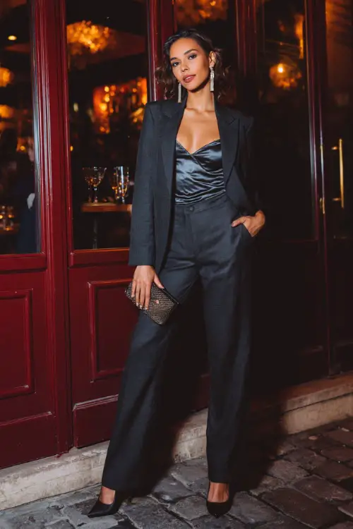 A woman dressed in a tailored blazer worn over a satin camisole, straight-leg trousers, and heeled boots, styled with a clutch and statement earrings