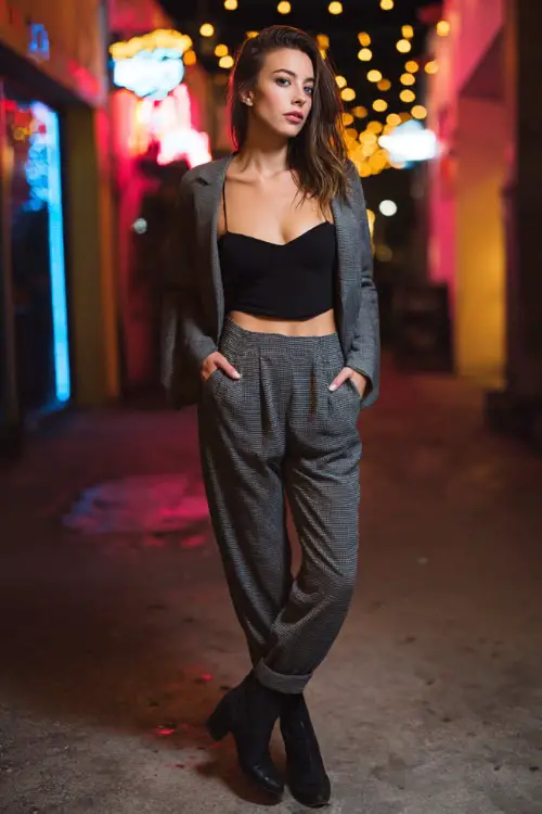 A woman wearing a knit top paired with tailored pants, layered with a relaxed blazer and ankle boots, standing in a casual winter nightlife setting 