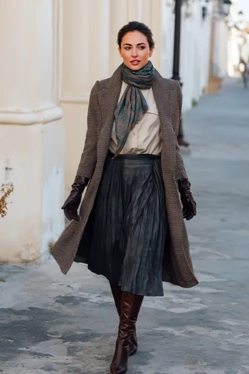 A woman wearing a sophisticated vintage scarf coat layered over a blouse and pleated skirt, styled with heeled boots and leather gloves
