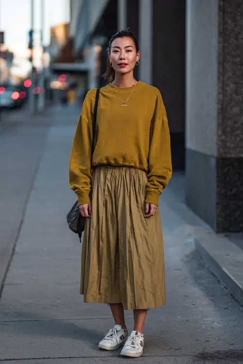 A woman wearing a vintage sweatshirt tucked into a pleated skirt, paired with sneakers and a casual shoulder bag, posed on a city sidewalk, full body framing