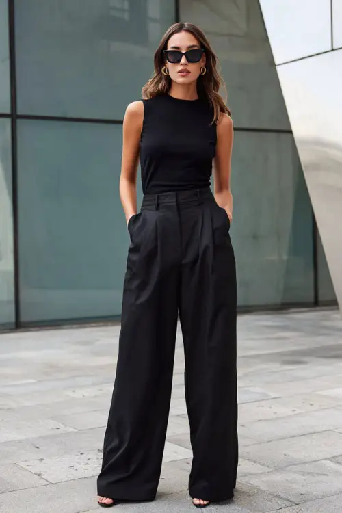 A woman wearing an all-black outfit with a sleeveless top and wide-leg trousers, styled with statement sunglasses and minimalist heels, standing in a contemporary urban plaza 