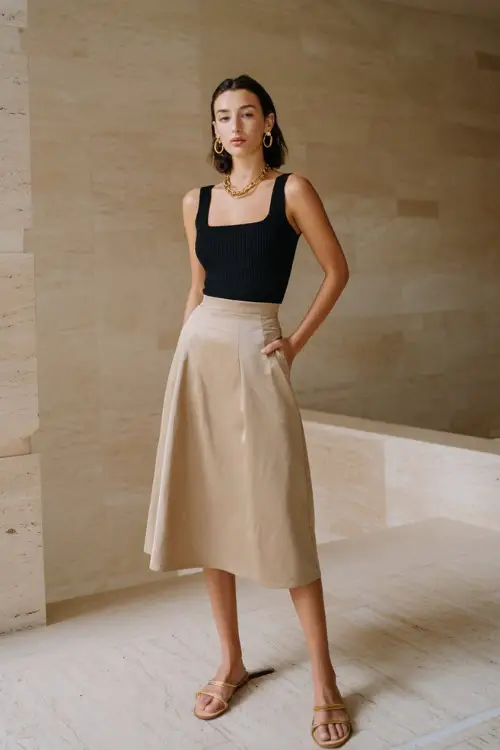 A woman wearing a black ribbed tank top paired with a neutral satin midi skirt, styled with minimalist sandals and delicate gold jewelry