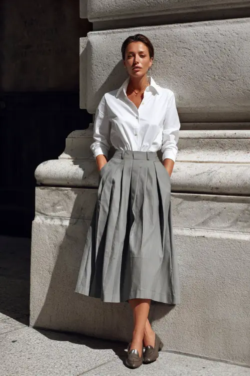 A woman wearing a crisp white button-down shirt tucked into a pleated gray midi skirt, styled with loafers and a slim belt 