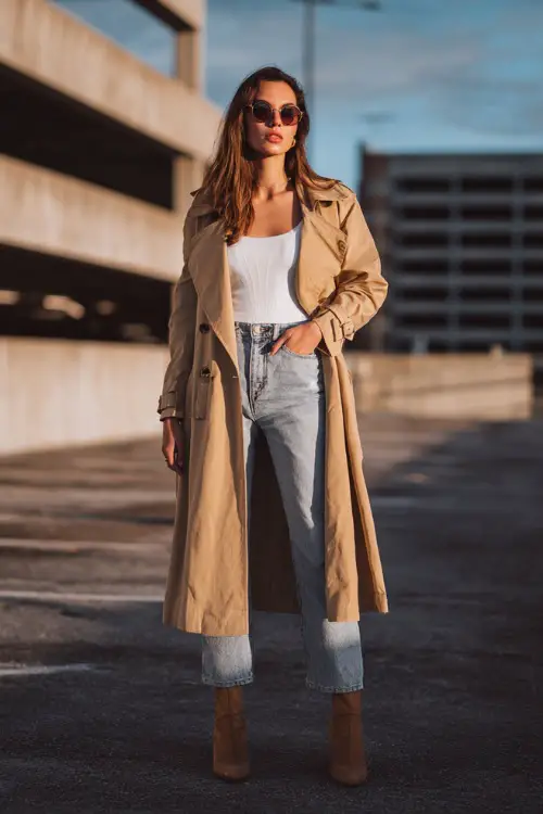 A woman wearing a lightweight camel trench coat layered over a white top and straight-leg jeans, styled with ankle boots and sunglasses