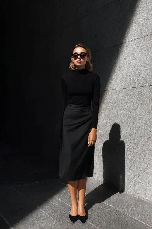 A woman wearing a monochrome all-black outfit with a fitted top and minimalist midi skirt, styled with pointed flats and sunglasses