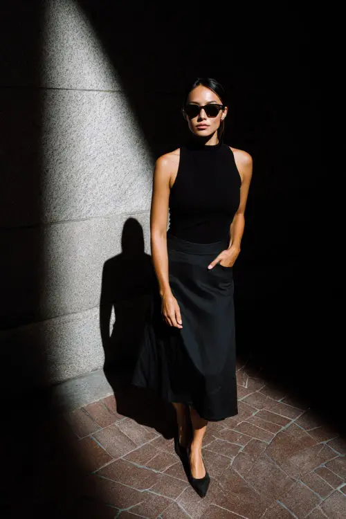 A woman wearing a monochrome all-black outfit with a fitted top and minimalist midi skirt, styled with pointed flats and sunglasses