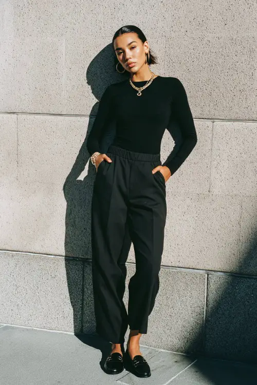 A woman wearing a monochrome black outfit with a fitted top and straight-leg pants, styled with simple loafers and subtle jewelry, photographed with minimal lighting 